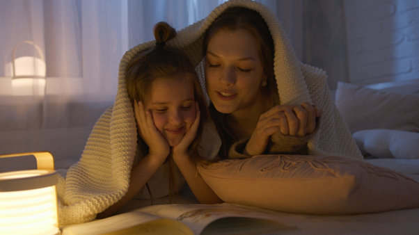 Mother and daughter happily reading a book under a white blanket, bonding and enjoying quality time together.