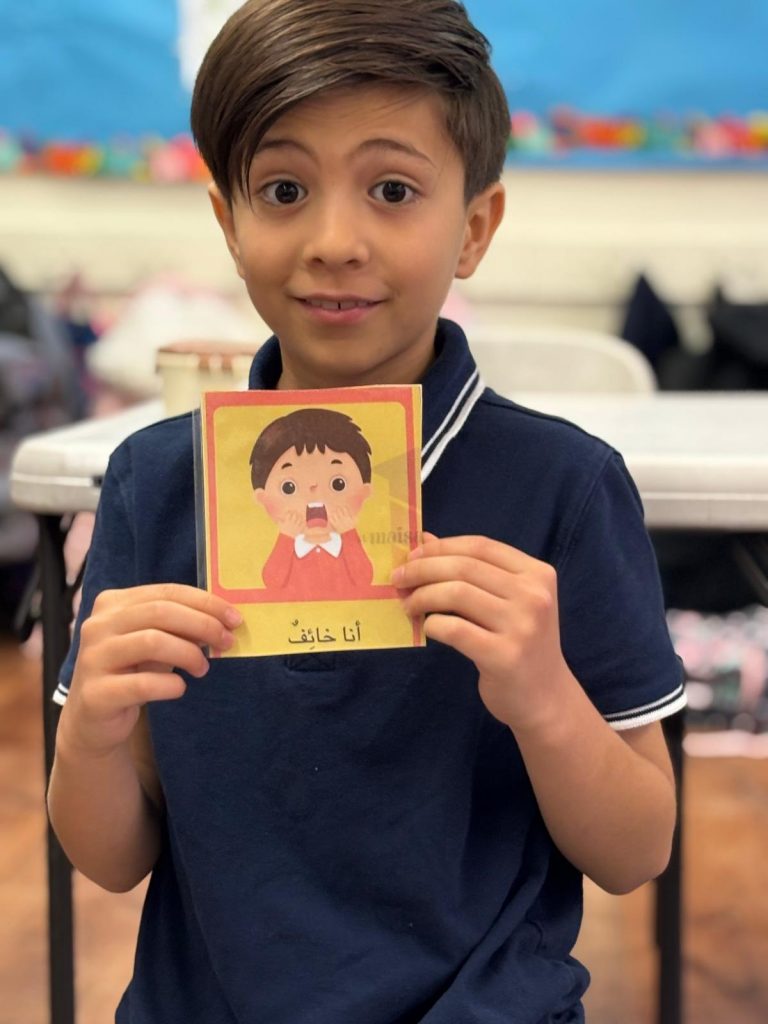 Boy in upper elementary holding a small picture that shows a cartoon boy who is scared and it says in arabic "I am scared"